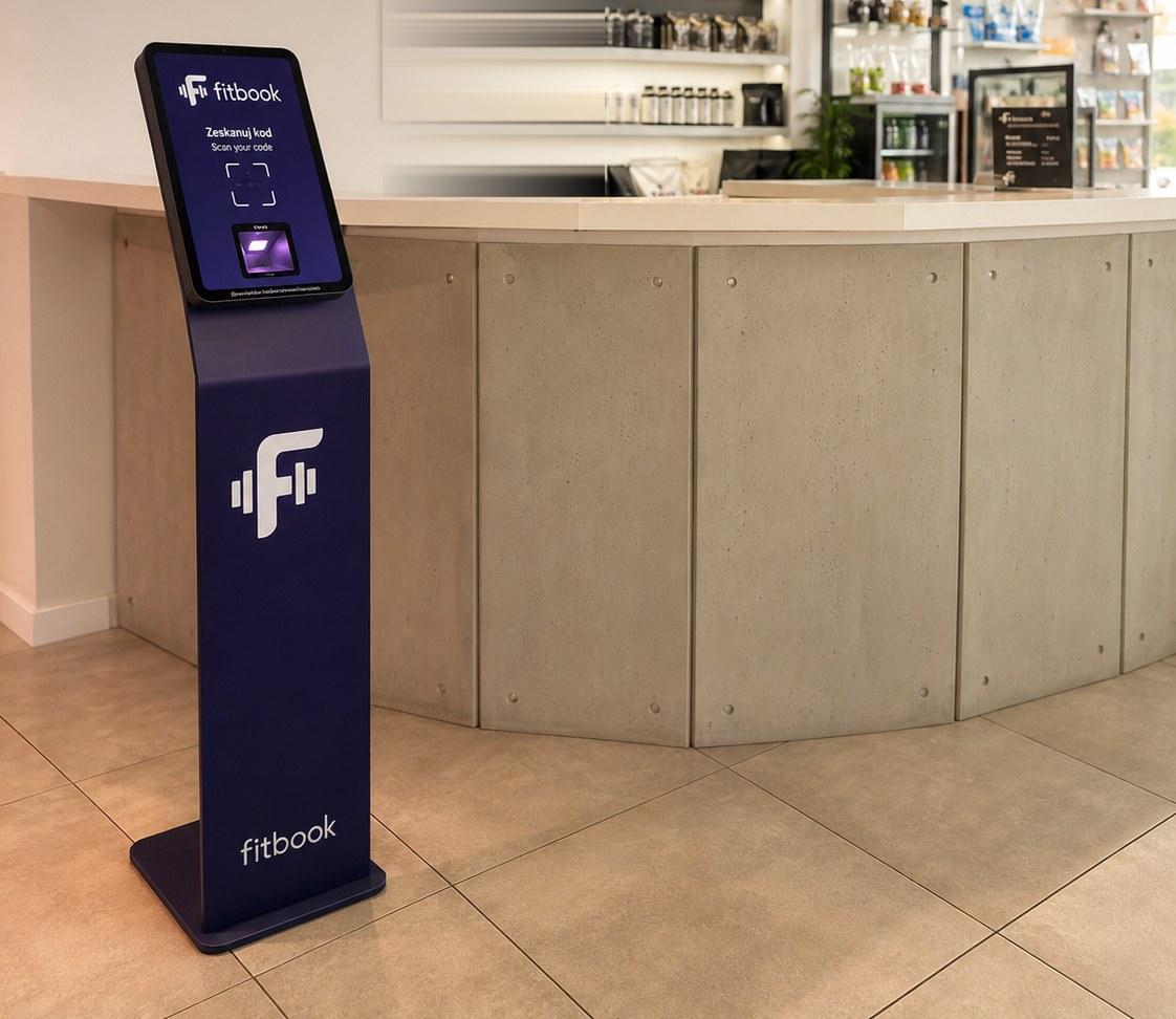 Kiosk FitBooka z ekranem „Zeskanuj kod” i czytnikiem, ustawiony przy ladzie recepcji siłowni